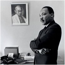 Black-and-white photo of a man in a suit standing with arms crossed in an office; a framed portrait of Gandhi hangs on the wall behind him.